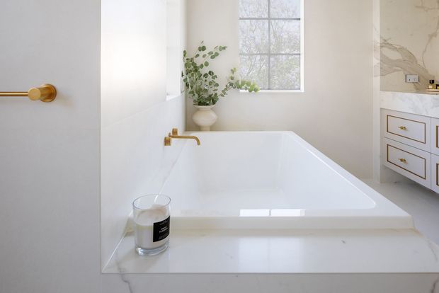 Bright, refined bathroom featuring a built-in white bathtub beneath a gridded window, with a white marble-look surround and walls. Brass/gold fittings, a potted plant, and a partial view of white vanity cabinetry with brass trim and hardware add warmth and detail.