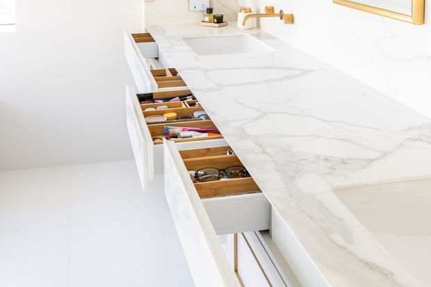 Bright, minimalist bathroom vanity viewed from above, featuring a long white stone countertop with grey veining, integrated basin, brushed gold wall-mounted tapware, and extensive white cabinetry with open drawers containing grooming items and accessories.