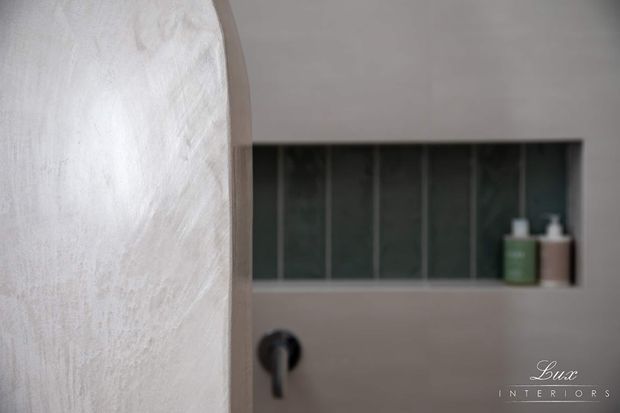 Close-up view of a refined bathroom interior featuring a curved plastered wall or partition in the foreground, a recessed wall niche lined with dark vertical tiles, and minimalist dark plumbing fixtures with green and neutral toiletry bottles. The palette is muted and contemporary with soft, textured finishes.