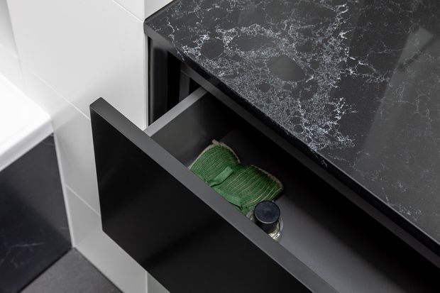 Close-up of a contemporary dark kitchen or bathroom-style drawer pulled open beneath a black stone-look benchtop with prominent white veining. The drawer front and cabinetry are matte black, with a small green fabric item and container inside. White tiled wall surfaces and dark flooring are visible around the joinery.