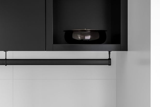 Close-up of a minimalist built-in kitchen or utility storage installation, featuring matte black overhead cabinetry with an open display niche and a black horizontal rail mounted against a white tiled wall. The composition is monochromatic and highly refined, with clean geometric lines and concealed detailing.