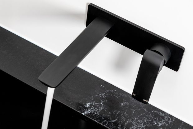 Close-up product image of a matte black wall-mounted bathroom tap set, with a rectangular backplate, angular spout, separate cylindrical control, and white outlet pipe above a dark countertop or basin edge. The minimalist fixture is photographed against a clean white background.