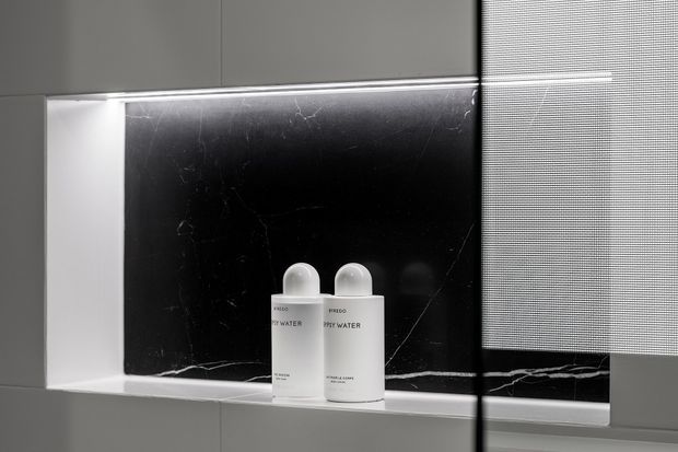 Close-up of a contemporary bathroom shower niche with a black marble-look back panel, pale tiled surround, and two white toiletry bottles. A partially lowered roller blind is visible at the right edge.