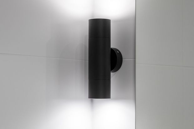 Close-up of a sleek black cylindrical wall-mounted light fixture on a white tiled wall. The fixture casts soft upward and downward illumination, creating a minimalist, high-contrast architectural detail.