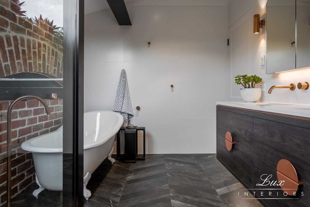 A refined bathroom with a freestanding white clawfoot bathtub, dark herringbone-patterned floor tiles, white tiled walls, and a dark timber vanity with a white countertop. Brass wall-mounted tapware, a large mirror, a black-framed glass shower enclosure, and exposed dark ceiling beams add contrast. A brick wall is visible through the glazing, and the space has a warm, contemporary-traditional feel.