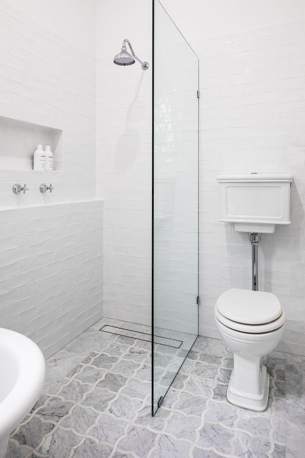 Bright, compact traditional-style bathroom featuring white glossy subway wall tiles, a frameless glass walk-in shower with chrome rain showerhead and controls, a wall-mounted cistern toilet, and patterned grey-and-white floor tiles. A partial freestanding bathtub is visible at the left edge.