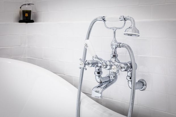Close-up of a traditional chrome bathtub faucet and shower assembly mounted against white rectangular wall tiles. A white freestanding bathtub curves into the foreground, with flexible shower hoses, crosshead controls, and a wall-mounted shower handset visible. A small dark soap dispenser sits on a wall shelf.