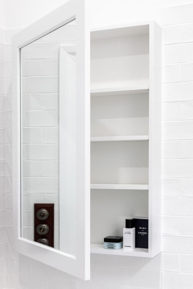 Bright white bathroom wall with glossy white subway-style tiles, a tall recessed open shelving niche containing toiletries, and a large white-framed mirrored cabinet door. The mirror reflects the tiled wall and a dark wall-mounted fixture. The space is clean and minimalist with strong white-on-white detailing.