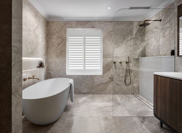 A refined contemporary bathroom featuring a freestanding white bathtub, walk-in shower with frameless glass screen, wall-mounted shower fittings, large marble-look wall and floor surfaces, white plantation shutters, white mosaic tile detailing, and a dark timber floating vanity.