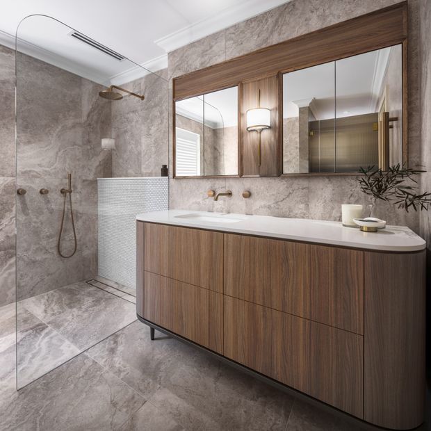 A refined contemporary bathroom featuring a large walk-in shower with frameless glass, grey stone-look wall and floor finishes, a wood-veneer floating vanity with integrated basin, and a wide framed mirrored cabinet with integrated lighting. Brushed brass tapware and shower fittings add warmth, while a freestanding towel radiator and minimal styling complete the space.