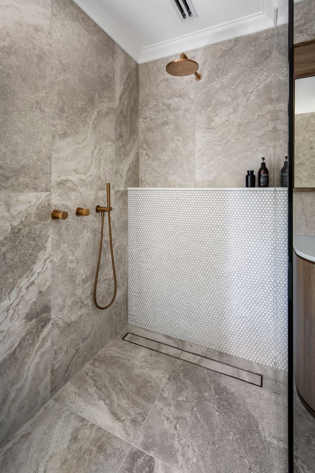 A refined walk-in shower with large-format grey-beige marble-look stone tiles covering the walls and floor. A white circular mosaic-tile half-height screen separates the shower from the vanity area, while brushed brass shower fittings and a rain shower add warmth. White decorative ceiling cornices and a mirrored vanity edge are visible.