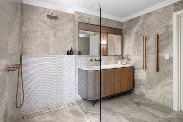 Contemporary bathroom with full-height marble-look wall and floor surfaces, a walk-in shower enclosed by a frameless curved glass screen, small-format mosaic shower walls, wall-mounted bronze shower fittings, and a floating timber vanity with integrated basin. A mirrored cabinet and warm vertical towel rails complete the refined fit-out.