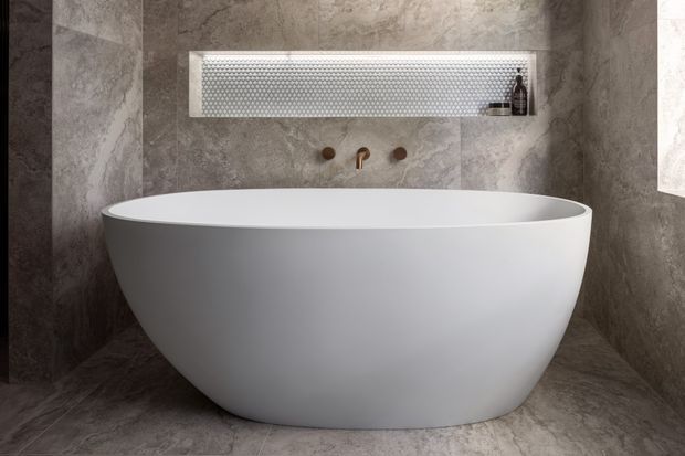 A contemporary bathroom featuring a freestanding white oval bathtub against grey stone-look wall and floor tiles. A recessed wall niche with a patterned metallic or tiled back provides storage above the tub, with wall-mounted bronze-toned fittings.