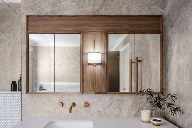 A refined contemporary bathroom vanity wall featuring a wide dark timber-framed mirrored cabinet, central brass wall sconce, pale stone-look wall finish, brass fixtures, and a light benchtop with a basin. The mirrors reflect a shower area and doorway, while a small plant and accessories add warmth.