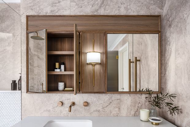 A refined bathroom vanity wall with a built-in dark timber mirrored cabinet, open shelving, integrated wall lighting, and brass-toned hardware. The cabinet is set against large light stone-look wall tiles, with a white basin and pale benchtop below. A freestanding-style tap set and small toiletries complete the calm, luxurious composition.