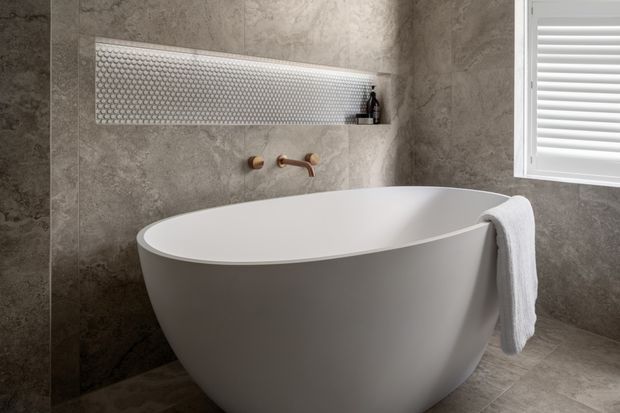 A refined contemporary bathroom featuring a freestanding white oval bathtub, large grey stone-look wall and floor tiles, a recessed shelf with small circular mosaic tiles, and brushed brass wall-mounted tapware. A white louvred window provides natural light.