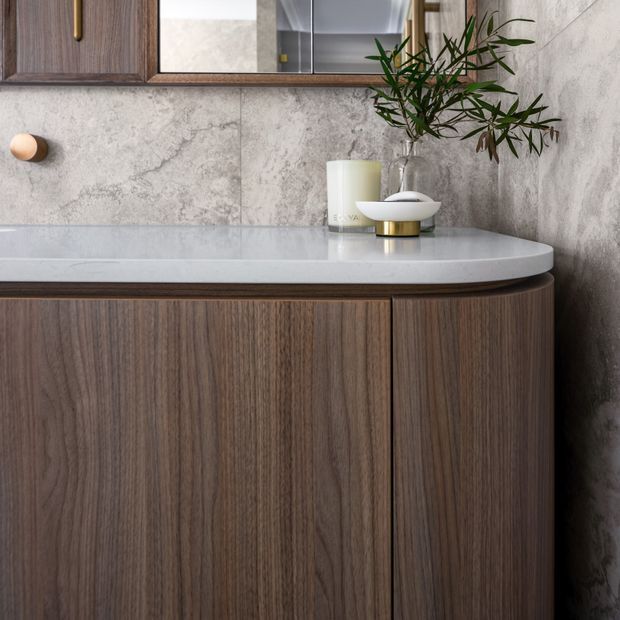 Close-up of a refined bathroom vanity featuring dark wood-grain cabinetry with rounded corners, a pale stone benchtop, large-format mottled grey stone wall tiles, and brass hardware. A mirrored cabinet and simple decorative accessories sit above the vanity, creating a warm, sophisticated finish.