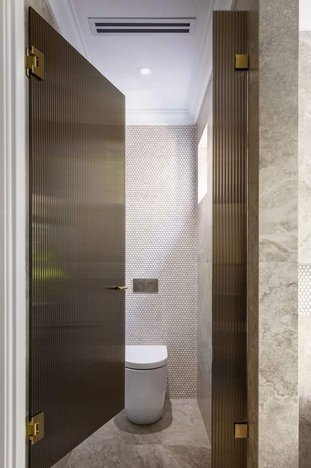A narrow, refined powder room viewed through an open fluted dark glass door. The compact space features a wall-hung toilet, small metallic flush plate, grey stone-look wall and floor finishes, and a decorative small-format circular mosaic tile feature wall. Brass-toned door hardware and warm dark fluted panels provide contrast, with recessed ceiling lighting and ventilation.