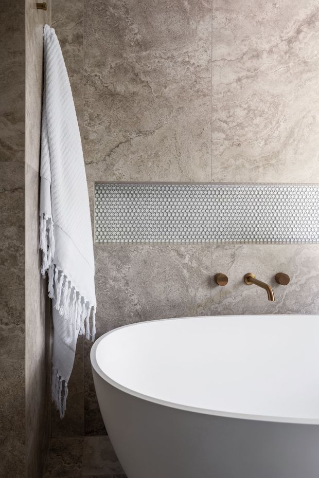 Close-up of a refined bathroom featuring a freestanding white oval bathtub against large grey-beige stone-look wall tiles. A horizontal band of small circular mosaic tiles adds a subtle feature detail, with brushed brass wall-mounted tapware and a white textured towel on a brass hook.