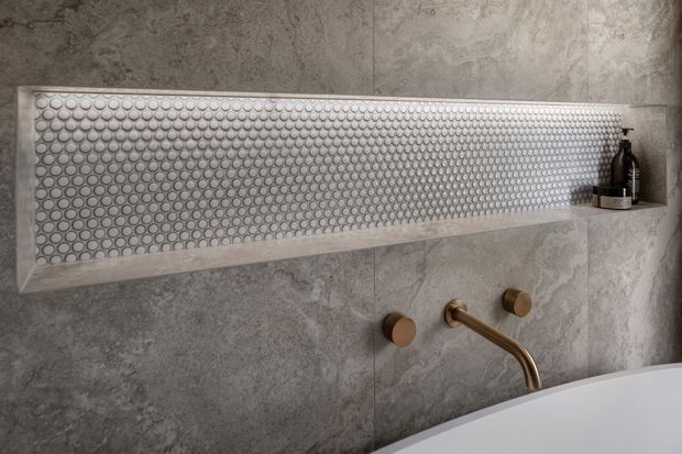 Close-up of a refined bathroom wall featuring a long recessed shelf lined with small circular mosaic tiles, set within large grey stone-look wall tiles. A brushed brass wall-mounted tap and matching controls sit above the edge of a white bathtub, with a dark soap dispenser on the shelf.
