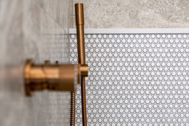 Close-up detail of a bathroom shower fixture in a warm brass or bronze finish, set against a small circular mosaic tile wall with pale grey grout and a textured grey stone-like surface.