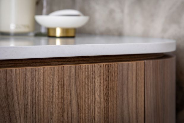 Close-up detail of a refined built-in timber vanity or cabinet with vertically grained dark wood cabinetry, a pale stone benchtop, and a textured stone wall finish. A small white and brass decorative object sits on the counter, emphasizing the warm contemporary material palette.