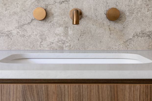 Close-up of a refined bathroom vanity featuring a white integrated rectangular basin with a slim raised rim, a wall-mounted bronze-toned spout and two round controls, a textured grey stone-look wall surface, and dark timber-grain cabinetry below.