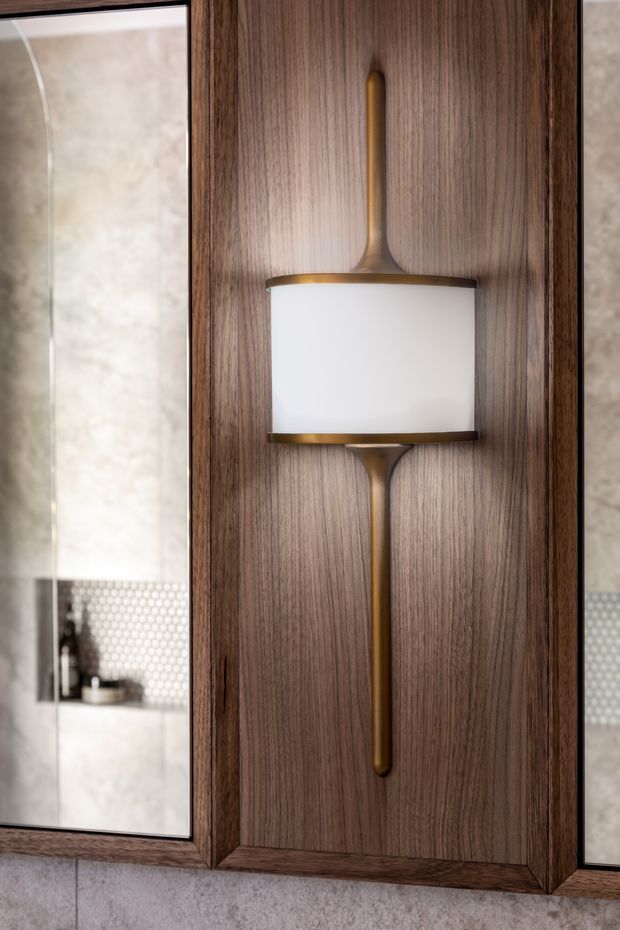 Close-up of a decorative wall sconce with a white rectangular diffuser and warm brass-toned vertical fittings, mounted on a rich dark timber veneer panel. Narrow mirror panels on either side reflect a bathroom vanity area with pale stone surfaces and small hexagonal mosaic tile detailing.