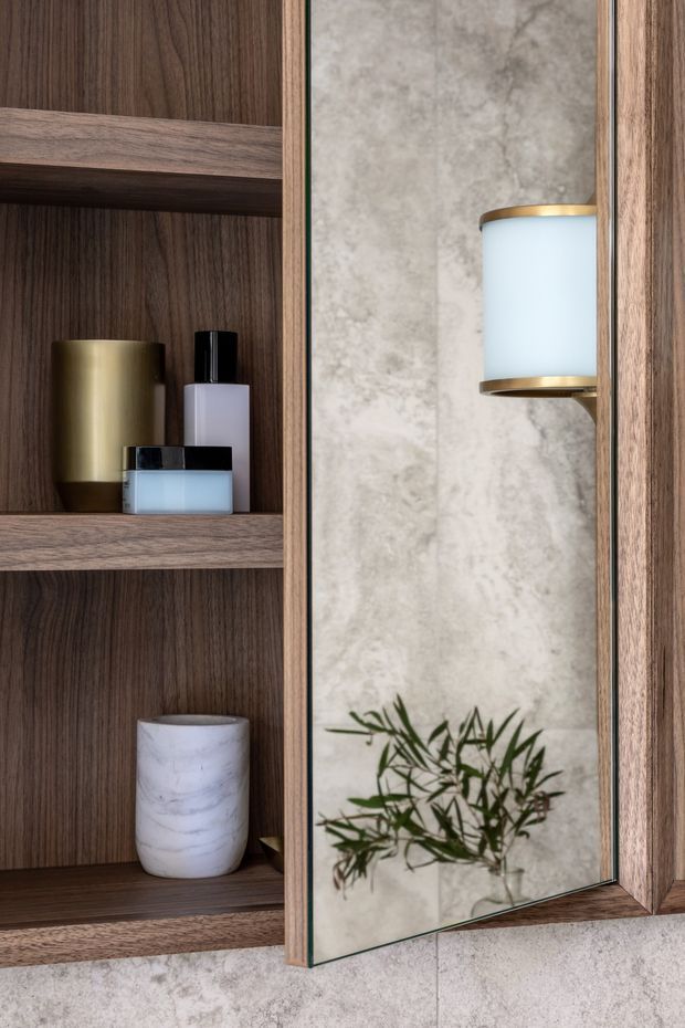 Close-up of a refined bathroom vanity or storage area with dark timber open shelving, a mirrored cabinet door, pale textured stone-look wall surfaces, and a brass-and-white wall sconce reflected in the mirror. Toiletries and a small plant add detail to the warm, modern joinery.