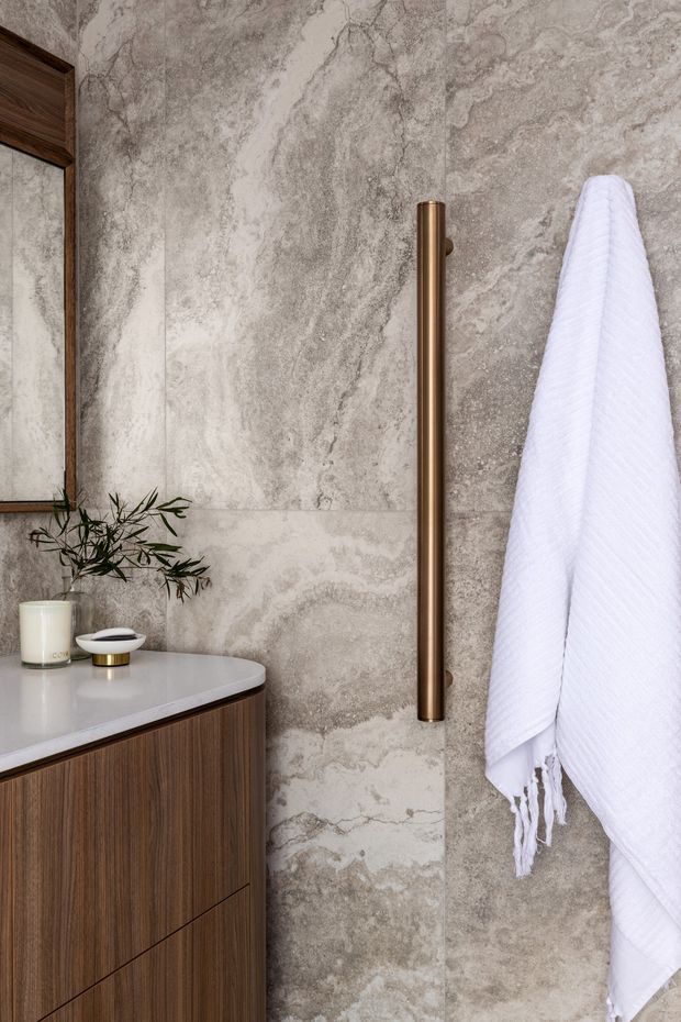 Close-up view of a refined bathroom vanity with dark timber cabinetry, a pale stone benchtop, large grey-beige veined stone wall tiles, and a vertical bronze towel rail. A frameless mirror, small basin accessories, a plant, and a white towel complete the warm contemporary scheme.