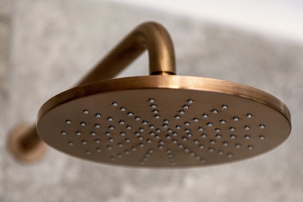 Close-up of a round copper-toned rain showerhead with many water outlets, mounted on a curved arm against a textured light-grey wall.
