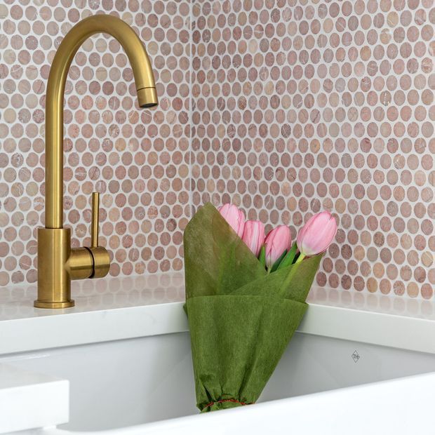 Close-up of a white inset bathtub or basin area with a tall brushed-gold gooseneck tap and matching control against a rounded pebble-pattern mosaic tile wall. A bouquet of pink tulips rests in the white fixture, adding a soft natural accent.