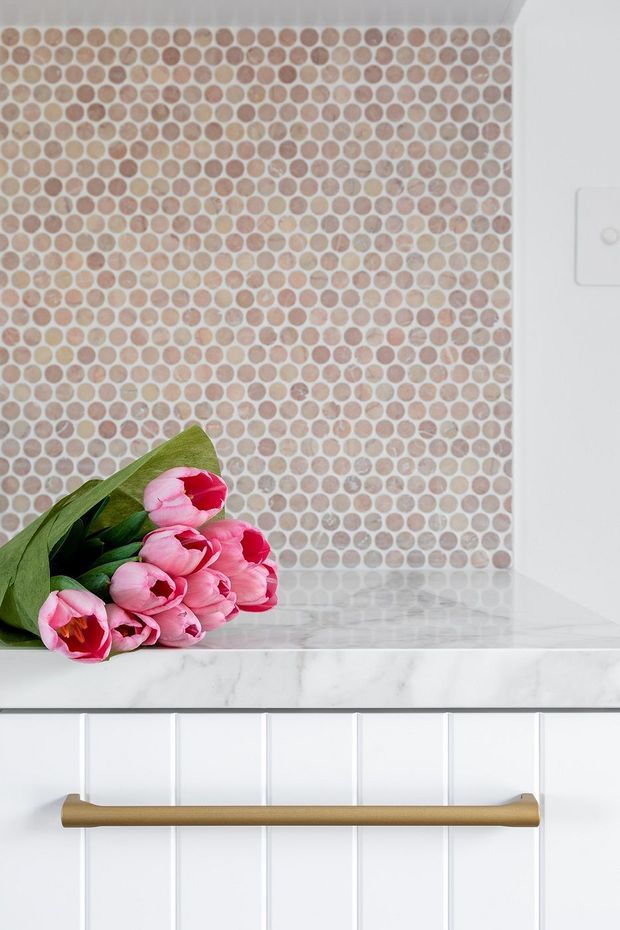 Close-up of a bright white built-in kitchen or utility-room cabinet featuring a polished white marble-look benchtop, a round blush and beige mosaic tile splashback, white vertical panelled cabinetry, and a long brushed brass-gold handle. Pink tulips rest on the benchtop as decorative styling.