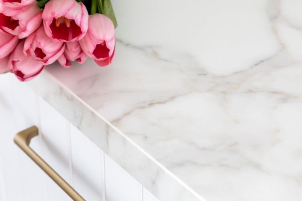 Close-up detail of a refined white kitchen or bathroom cabinet with a thick white marble-look countertop featuring soft grey veining and a long brushed brass-gold handle. Pink flowers sit on the benchtop as decorative styling. The image highlights the benchtop and cabinetry rather than the full room.