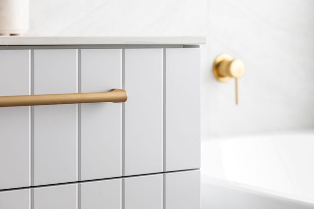 Close-up detail of a refined bathroom vanity or built-in cabinet with vertically fluted light-grey cabinetry, a slim pale countertop, and a warm brass cylindrical handle. A matching brass wall control is visible beside a white bathtub, creating a clean, softly lit contemporary bathroom setting.
