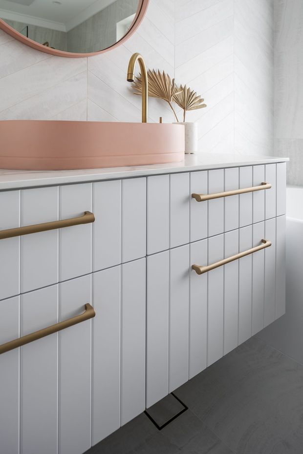 A close-up view of a contemporary bathroom vanity with pale grey vertical-panel cabinetry, a white stone benchtop, a rounded blush-pink vessel basin, and a brushed brass tap and handles. White geometric wall tiles, a large round mirror with a pink-toned frame, dried palm leaves, and dark grey floor tiles complete the softly styled space.