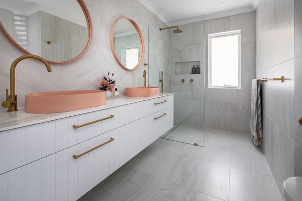 Contemporary bathroom with a long white vertically panelled vanity, twin blush-pink vessel basins, brushed brass tapware and hardware, round copper-toned framed mirrors, and large pale grey marble-look wall and floor tiles. A frameless glass shower, recessed niche, window, heated towel rail, and freestanding bath edge are visible. The palette is light, warm, and refined.