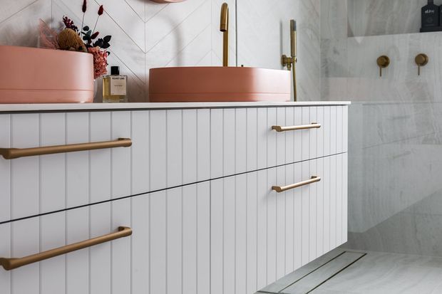 Close-up view of a contemporary bathroom vanity with vertically fluted white cabinetry, a pale pink vessel basin, brushed brass tapware and handles, and large light grey marble-look wall and floor tiles. A wall-mounted shower fixture is visible in the background.