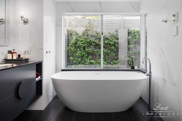 Contemporary bathroom featuring a freestanding white oval bathtub beneath a large opening window, a wall-mounted dark vanity, marble-look wall finishes, black floor tiles, chrome fixtures, and a leafy courtyard visible outside.