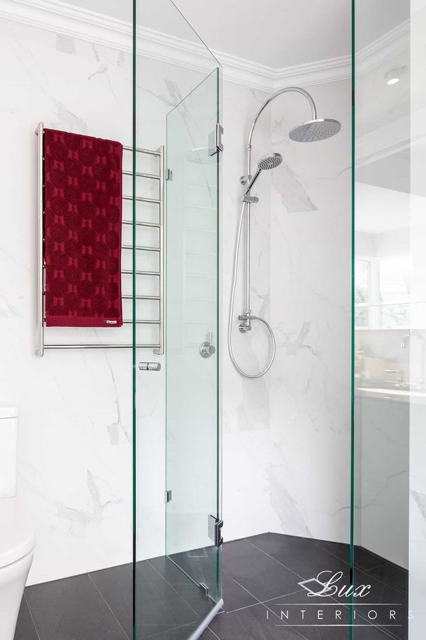 Bright contemporary bathroom featuring a frameless glass walk-in shower with dual chrome showerheads, white marble-look wall tiles, dark charcoal floor tiles, and a chrome heated towel rail holding a deep red towel. A freestanding or partially visible white toilet and vanity area are seen at the edges and through the glass.