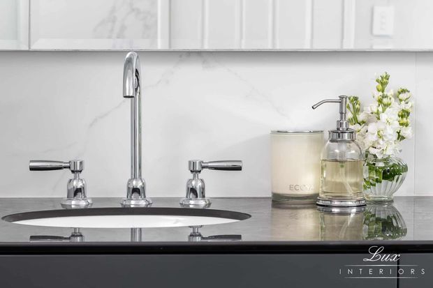 Close-up of a refined bathroom vanity with a polished chrome high-arc faucet, separate cross-handled taps, undermount basin, and dark reflective stone countertop. White marble-look wall tiling and cabinetry form the backdrop, with coordinated glass dispensers, a candle, and a vase of white flowers. Lux Interiors branding appears at the lower right.