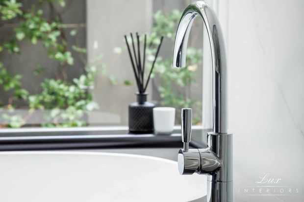 Close-up interior detail of a contemporary bathroom basin with a tall curved chrome mixer tap and separate cylindrical control. The white basin, dark window sill, neutral wall surfaces, and reflected greenery create a clean, refined setting.