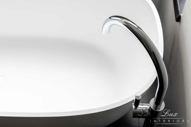 Close-up product-style image of a white freestanding bathtub with a sleek curved chrome floor-mounted faucet and matching controls. The composition is minimal and monochromatic, with the Lux Interiors logo visible in the lower-right corner.