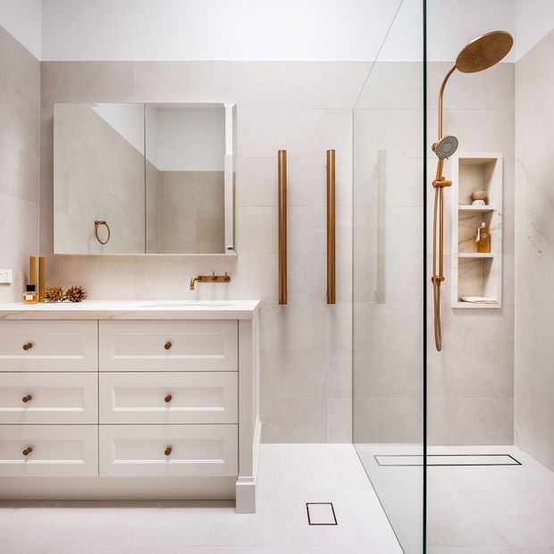 A bright, refined bathroom featuring a white panelled vanity with timber knobs, a pale stone-look tiled floor and walls, a large mirrored cabinet, brushed bronze wall-mounted tapware and grab rails, and a frameless glass walk-in shower with a bronze rain shower and recessed shelving.