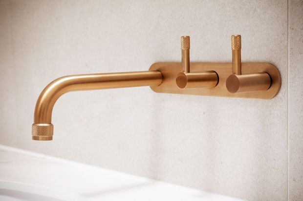 Close-up of a wall-mounted brushed brass or bronze bathtub tap set, with a curved spout and two cylindrical controls, installed against a light neutral tiled or plastered wall above a white bathtub.