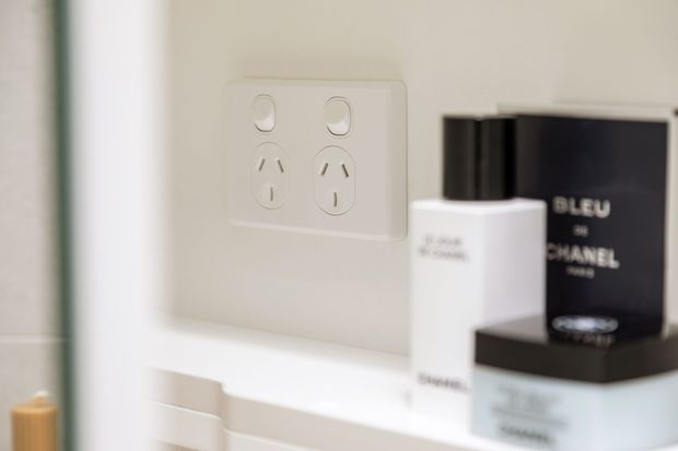 Close-up detail of a light neutral bathroom or vanity wall, featuring a white double power outlet above a white ledge or built-in surface. Minimal toiletry and fragrance containers sit in the foreground, with a softly blurred mirror or vertical edge at left.