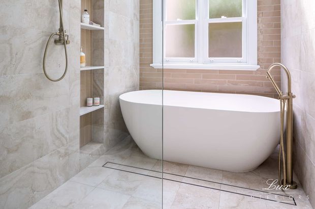 Bright, refined bathroom featuring a freestanding white oval bathtub beneath a window, a frameless glass shower screen, wall-mounted shower fittings, recessed open shelving, pale stone-look wall and floor tiles, and brushed brass tapware.