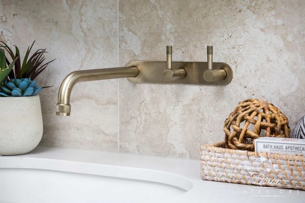 Close-up of a refined bathroom or ensuite bathtub area featuring a brushed brass wall-mounted bath mixer with a curved spout and twin controls, set against large-format beige natural-stone-look wall tiles. A white freestanding or built-in bath edge, potted plants, and woven baskets add warm, organic styling.
