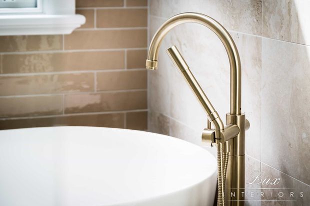 Close-up of a freestanding bathtub with a tall brushed brass/gold floor-mounted faucet and handheld shower attachment. The bath is surrounded by beige and cream tiled walls, with a small white-framed window visible at upper left. The image focuses on refined bathroom fixtures and finishes.