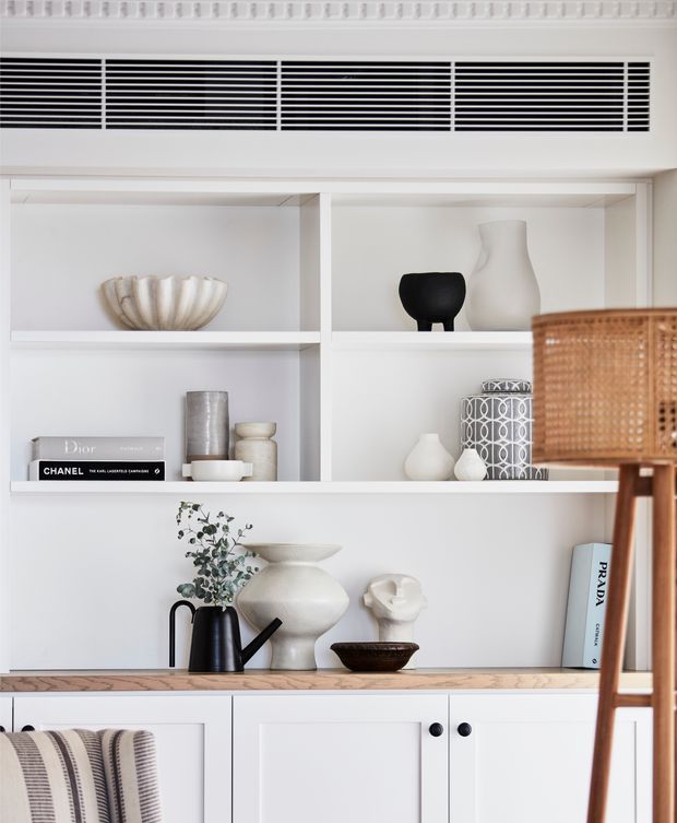 Close-up of a refined white built-in shelving and cabinetry wall, styled with neutral ceramic and glass vessels, art books, a sculptural bust, and a small plant. A woven rattan pendant or furniture element occupies the right foreground, while a striped textile appears at lower left. The composition is bright, minimalist, and softly tonal, with decorative ceiling trim and a long horizontal ventilation grille above the shelving.